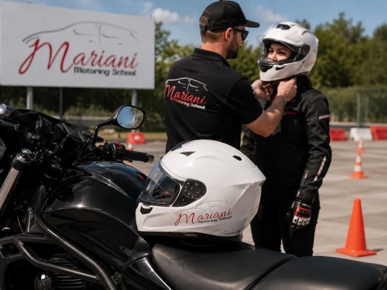 A Mariani instructor helping a learner fasten a white Mariani-branded helmet beside the training motorcycle